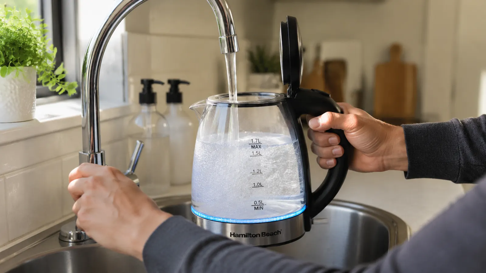 Hand filling a Hamilton Beach electric kettle at a sink with a small kitchen visible in the background