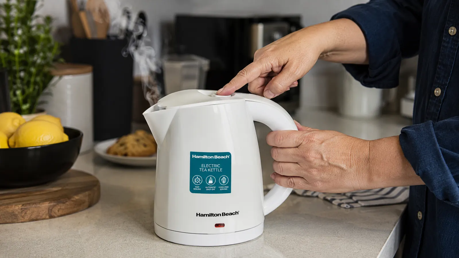 Hand pressing the power button on a Hamilton Beach 1-liter electric kettle