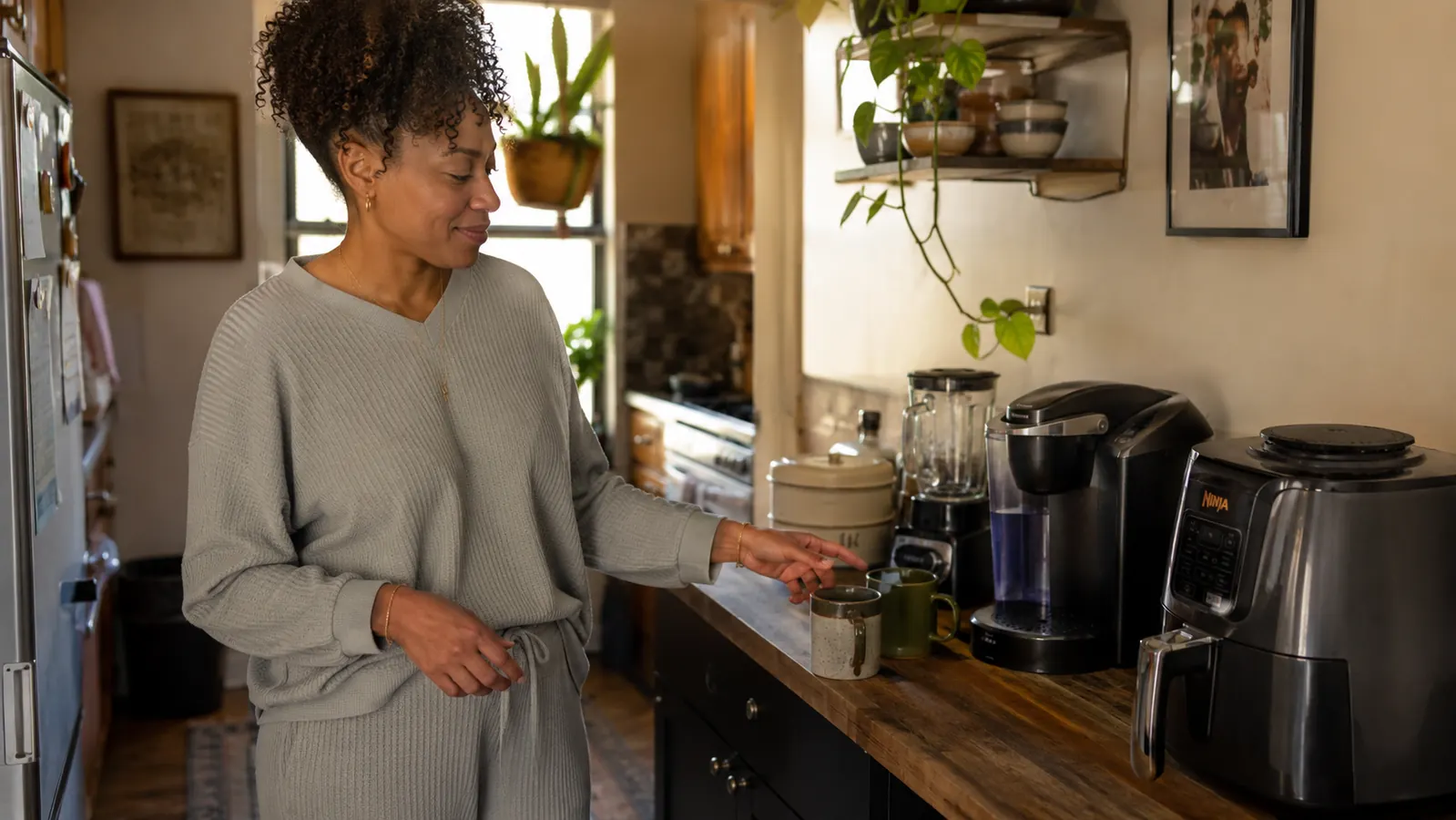 A woman in her fifties at a sunlit kitchen window with coffee and a journal, calm morning light